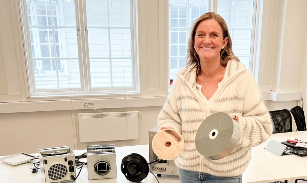Monica Aarethun holder en liten og en stor avfukterrotor, med tre avfukter på et bord i bakgrunnen,