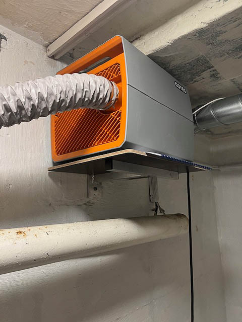 En Cotes CL26 avfukter plassert på stativ høyt på veggen, med fleksibel kanal for avkastluft ut fra baksiden av avfukteren,
